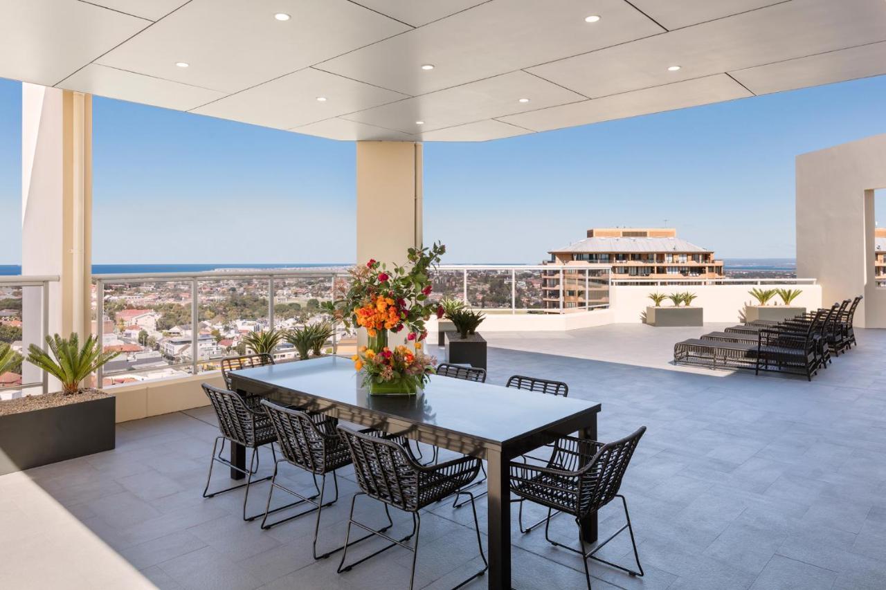 Meriton Suites Bondi Junction Sydney Kültér fotó The photo shows a stylish outdoor terrace or balcony area, featuring a long dining table surrounded by several black wire chairs. The table has a centerpiece of vibrant flowers. The space is well-lit, with a clear view of the horizon in the backgroun