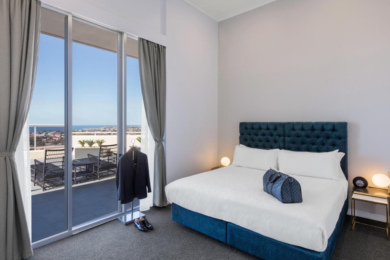 Meriton Suites Bondi Junction Sydney Kültér fotó The photo shows a stylish bedroom featuring a large bed with a tufted blue headboard. There's a neatly organized outfit consisting of a suit hanging on a stand, along with shoes placed on the floor. The room has large windows that provide a view of t