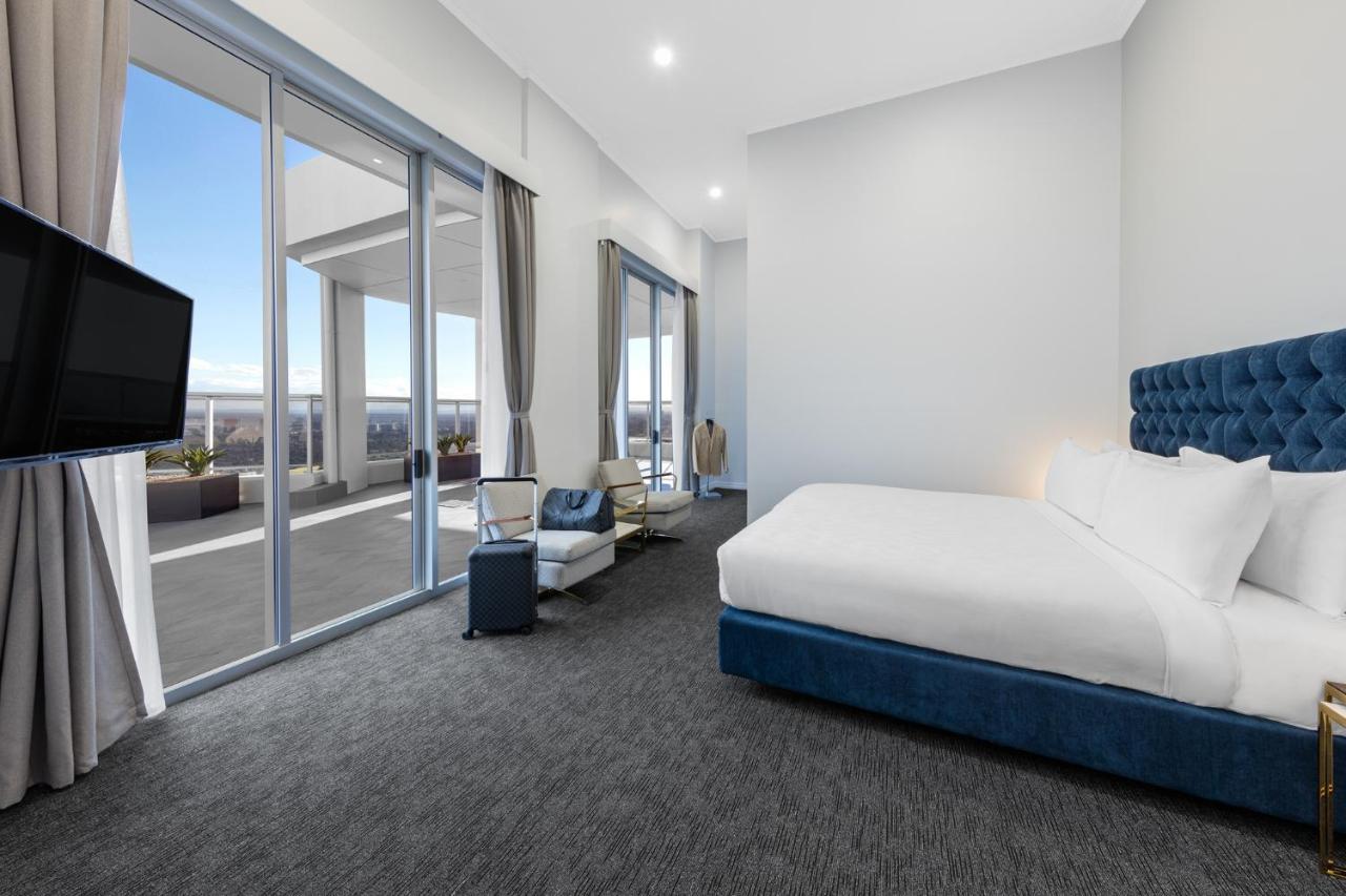Meriton Suites Bondi Junction Sydney Kültér fotó The photo shows a modern and spacious hotel room. It features a large bed with a blue headboard, a flat-screen TV mounted on the wall, and a cozy seating area with a chair and small table. The room has large windows that offer natural light and a vie