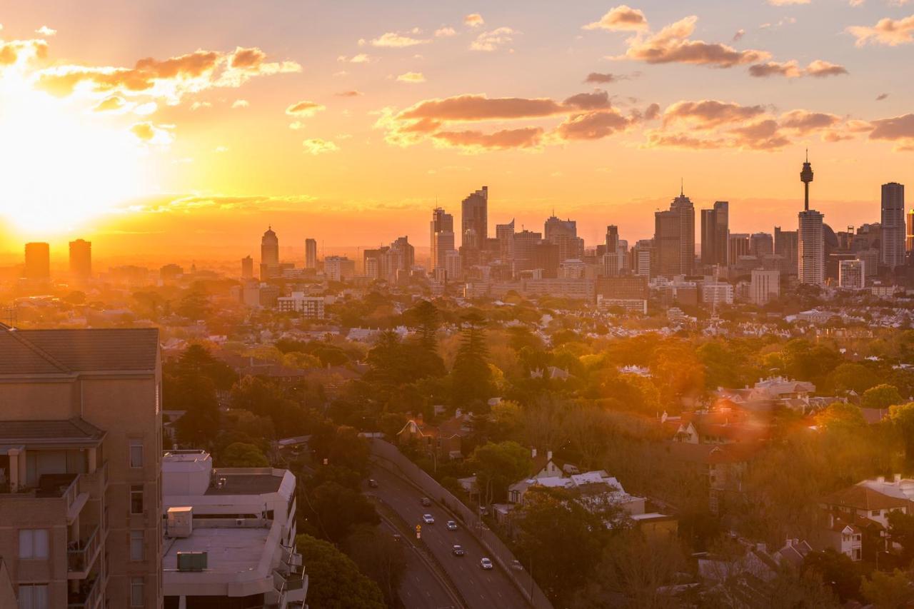 Meriton Suites Bondi Junction Sydney Kültér fotó The photo shows a panoramic view of a city skyline at sunset. The sun is low on the horizon, casting a warm golden light across the buildings. Tall skyscrapers are silhouetted against the colorful sky, which features hues of orange, pink, and yellow,