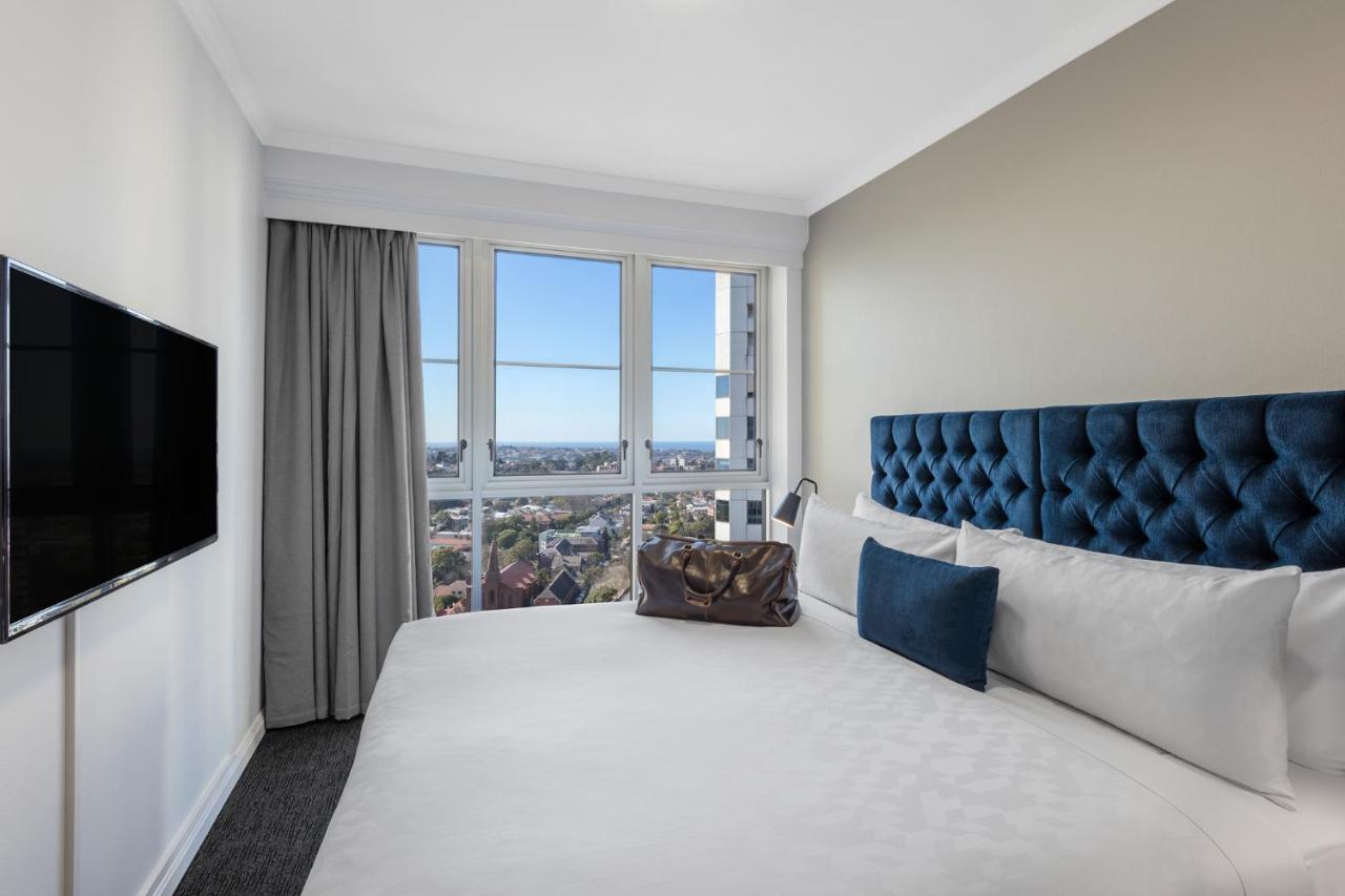 Meriton Suites Bondi Junction Sydney Kültér fotó The photo shows a modern hotel room featuring a large bed with a tufted blue headboard. Beside the bed, there is a stylish bag placed on the bed. The room has large windows that provide a view of the outside, allowing natural light to fill the space.