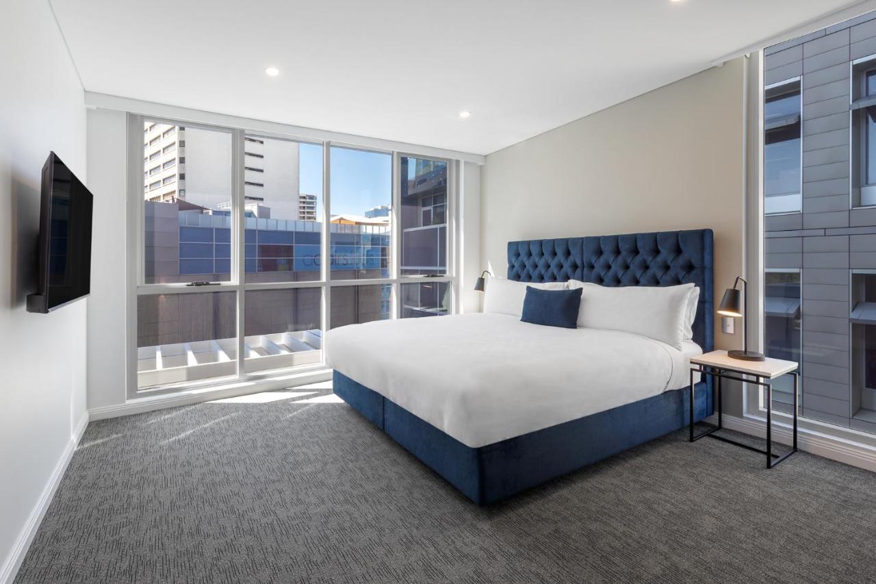 Meriton Suites Bondi Junction Sydney Kültér fotó The image shows a modern hotel room with a large, comfortable bed featuring a blue upholstered headboard. The room has large windows that allow natural light to enter, providing a good view of the surrounding buildings. There is a small bedside table