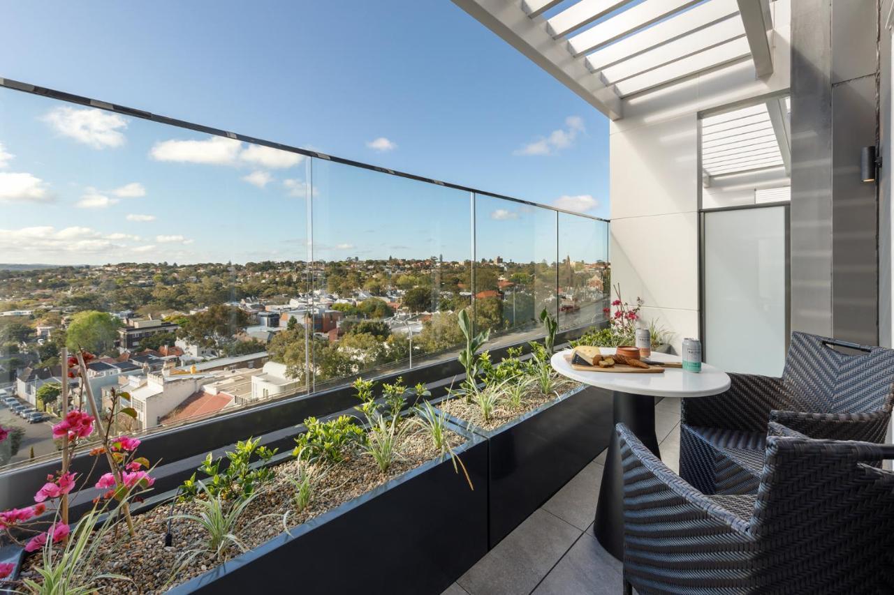 Meriton Suites Bondi Junction Sydney Kültér fotó The photo shows a modern balcony with a glass railing, enhancing the view of the surrounding area, which includes various rooftops and greenery in the distance. There is a small round table set with a decorative floral arrangement and some items, lik