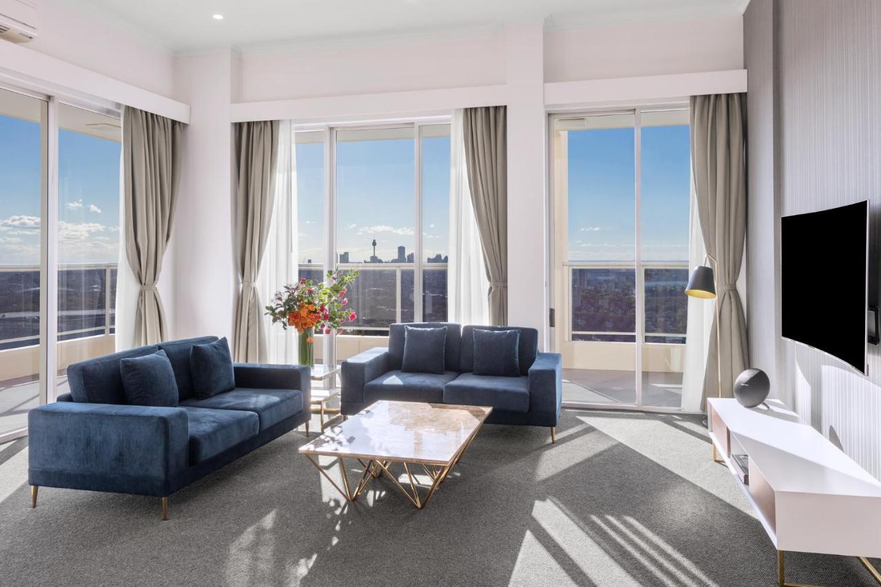Meriton Suites Bondi Junction Sydney Kültér fotó The photo shows a modern living room with large windows that allow natural light to fill the space. There are two blue sofas arranged around a coffee table with a glass top and gold legs. A floral arrangement adds a touch of color, placed on a nearby