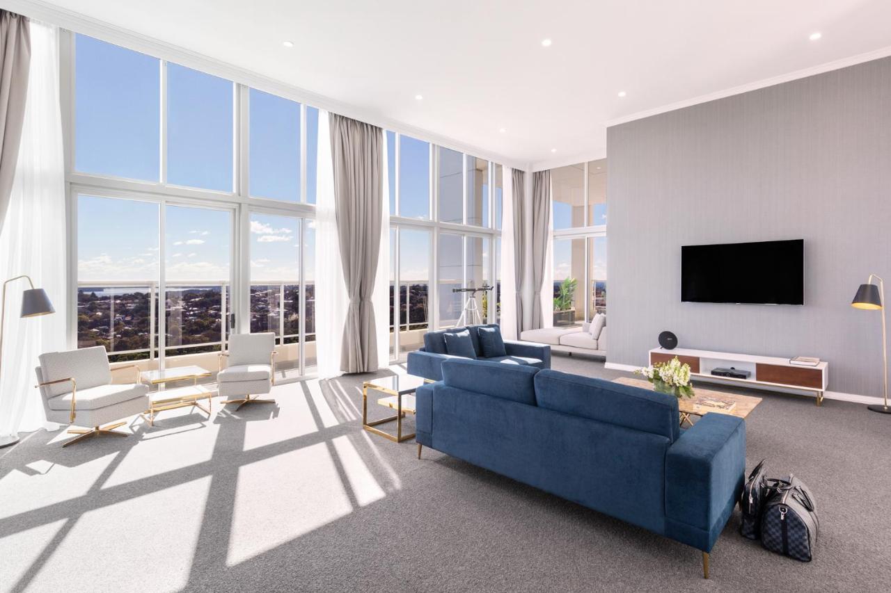 Meriton Suites Bondi Junction Sydney Kültér fotó The photo shows a bright and modern living room with large floor-to-ceiling windows that provide a view of the outdoors. The space features a blue sofa in the center, complemented by light-colored armchairs nearby. There are light curtains hanging by