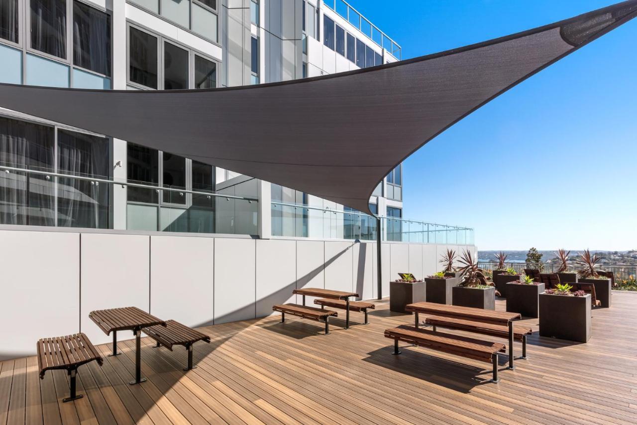 Meriton Suites Bondi Junction Sydney Kültér fotó The photo shows a modern outdoor terrace area on a building. It features several wooden benches arranged on a wooden deck. There are large planters with greenery, adding some decorative elements. A large triangular gray shade sail is positioned above