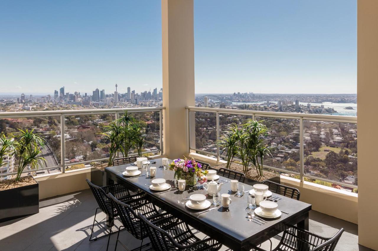 Meriton Suites Bondi Junction Sydney Kültér fotó The photo shows a beautifully arranged outdoor dining area on a balcony. There is a long black dining table set for a meal, with plates, glasses, and decorative items like a vase of flowers. Surrounding the table are stylish black chairs. In the back