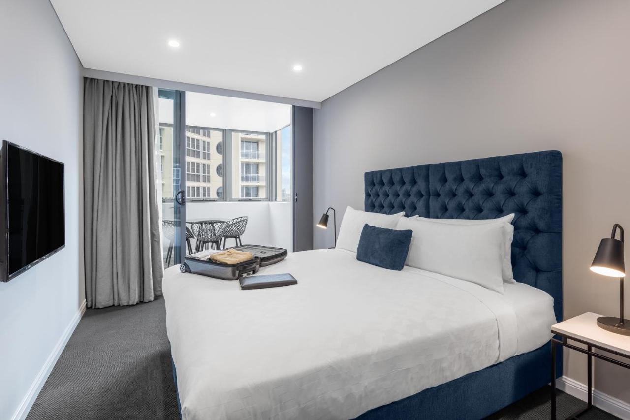 Meriton Suites Bondi Junction Sydney Kültér fotó The photo shows a modern hotel room featuring a large bed with a tufted blue headboard. There are minimalistic bedside tables with lamps on either side of the bed. A flat-screen TV is mounted on the wall across from the bed. To one side, there are fl