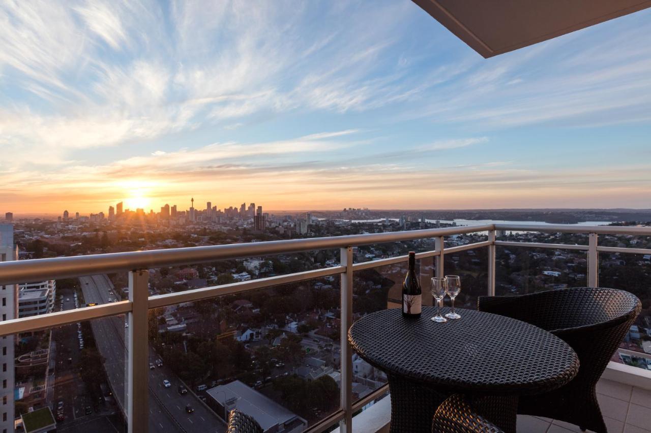 Meriton Suites Bondi Junction Sydney Kültér fotó The image shows a scenic view from a balcony or terrace overlooking a cityscape during sunset. In the foreground, there is a small round table with two glasses and a bottle of wine, suggesting a romantic or relaxing setting. The sky is filled with vi