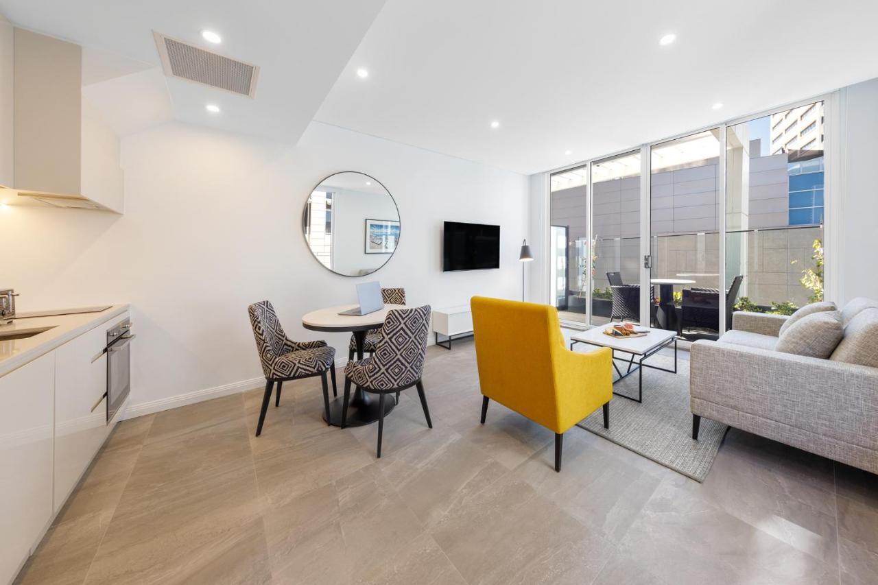 Meriton Suites Bondi Junction Sydney Kültér fotó The photo shows a modern and stylish living space. On one side, there is a kitchen area with sleek cabinetry, and next to it, a round dining table surrounded by three patterned chairs. In the center of the room, there is a gray sofa accompanied by a 