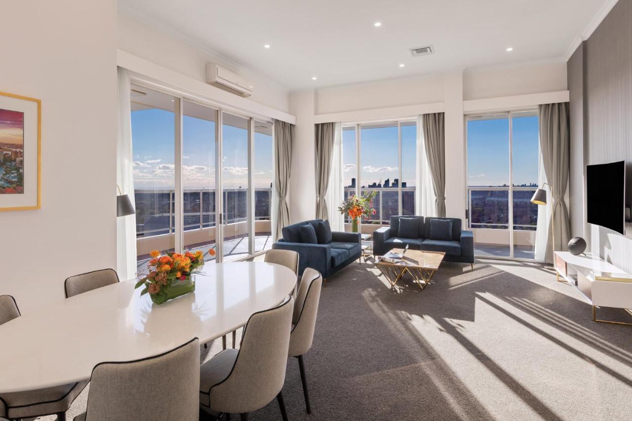 Meriton Suites Bondi Junction Sydney Kültér fotó The photo shows a modern and stylish living space with large windows allowing natural light to fill the room. In the foreground, there is a round dining table with several chairs around it, adorned with a floral centerpiece. The seating area features