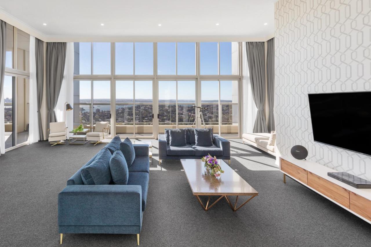 Meriton Suites Bondi Junction Sydney Kültér fotó The photo shows a modern and spacious living area. It features large windows that provide a panoramic view of the outside landscape and allow natural light to fill the room. The interior design includes a blue sectional sofa placed in the center, com