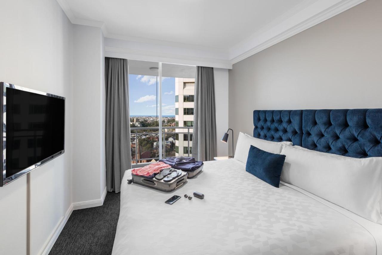 Meriton Suites Bondi Junction Sydney Kültér fotó The photo shows a modern hotel room featuring a neatly made bed with a blue tufted headboard and a white duvet. There's a small decorative pillow on the bed, and a piece of luggage is placed at the foot with clothes partially unpacked. A television i