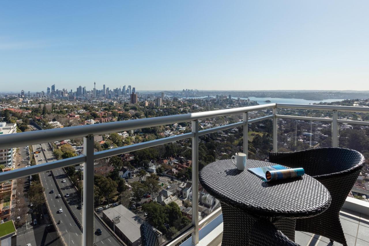 Meriton Suites Bondi Junction Sydney Kültér fotó The photo shows a balcony with a view overlooking a cityscape and a body of water in the distance. There is a round table with a woven design and two chairs, one of which has a mug on it. On the table, there’s also a rolled-up magazine or document. T