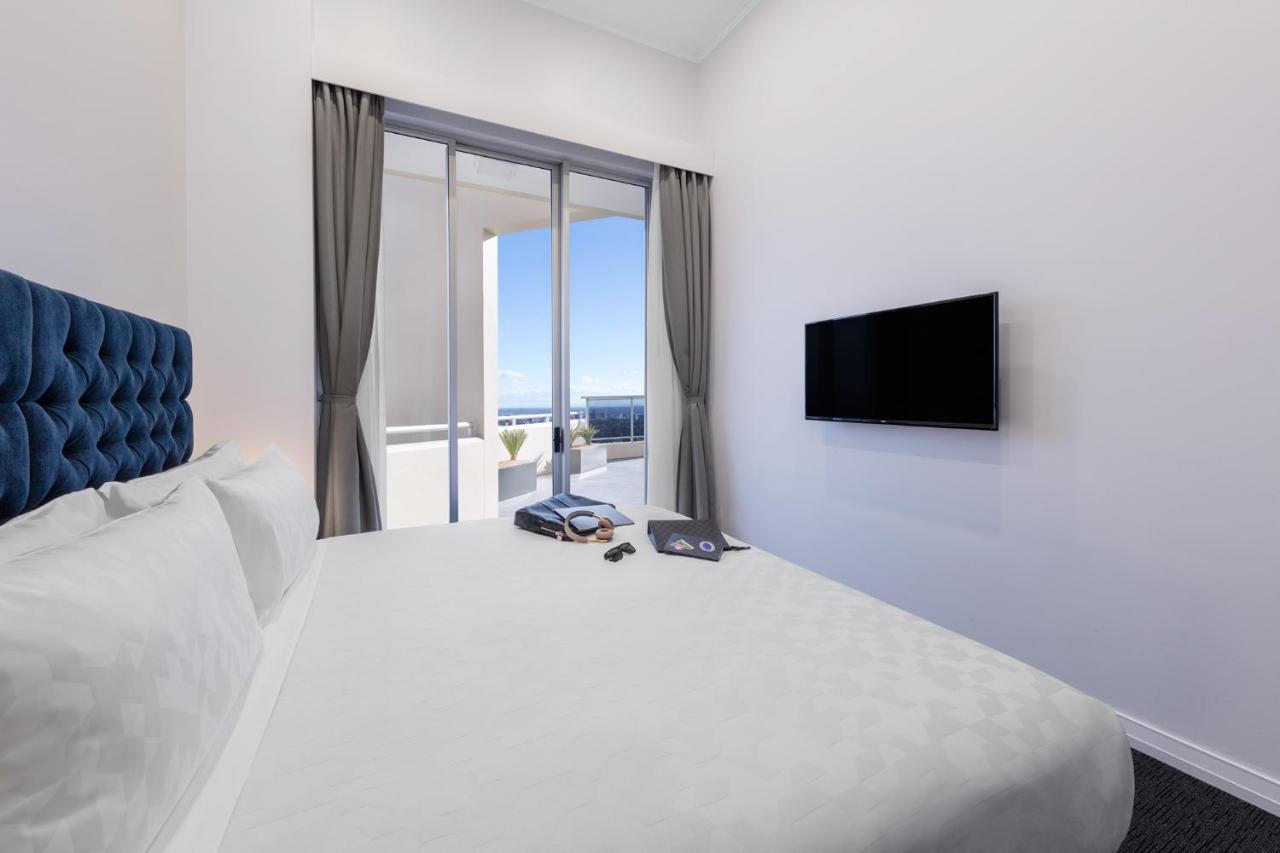 Meriton Suites Bondi Junction Sydney Kültér fotó The photo shows a modern hotel room with a large, comfortable bed featuring a blue tufted headboard. There are light-colored walls, and the room is decorated simply. A flat-screen TV is mounted on the wall opposite the bed. The room has large glass d