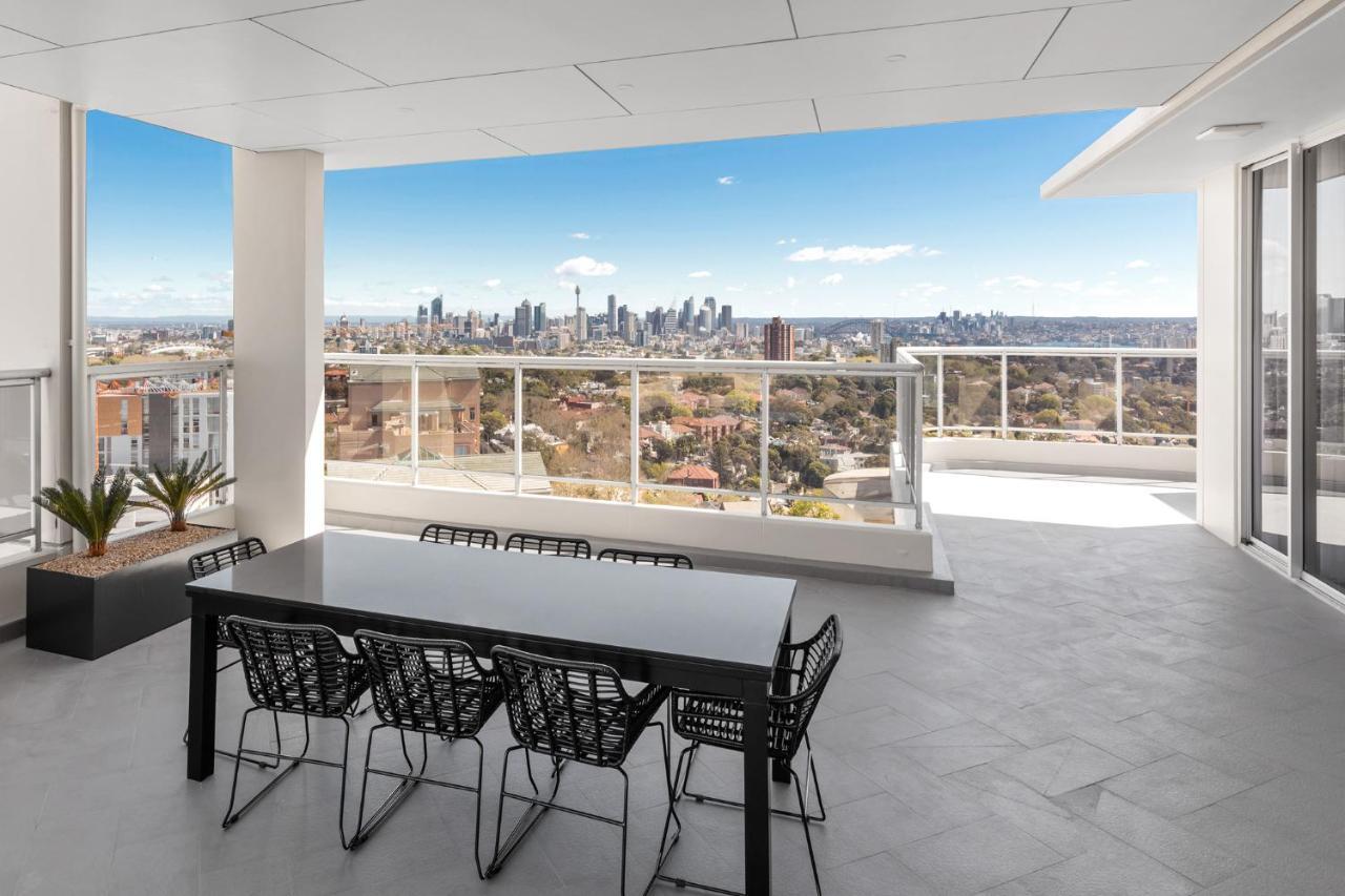 Meriton Suites Bondi Junction Sydney Kültér fotó The photo shows a modern outdoor balcony or terrace space with a dining table and chairs. The table is black and has a minimalist design, surrounded by stylish chairs. In the background, there is a stunning view of a city skyline under a clear blue s