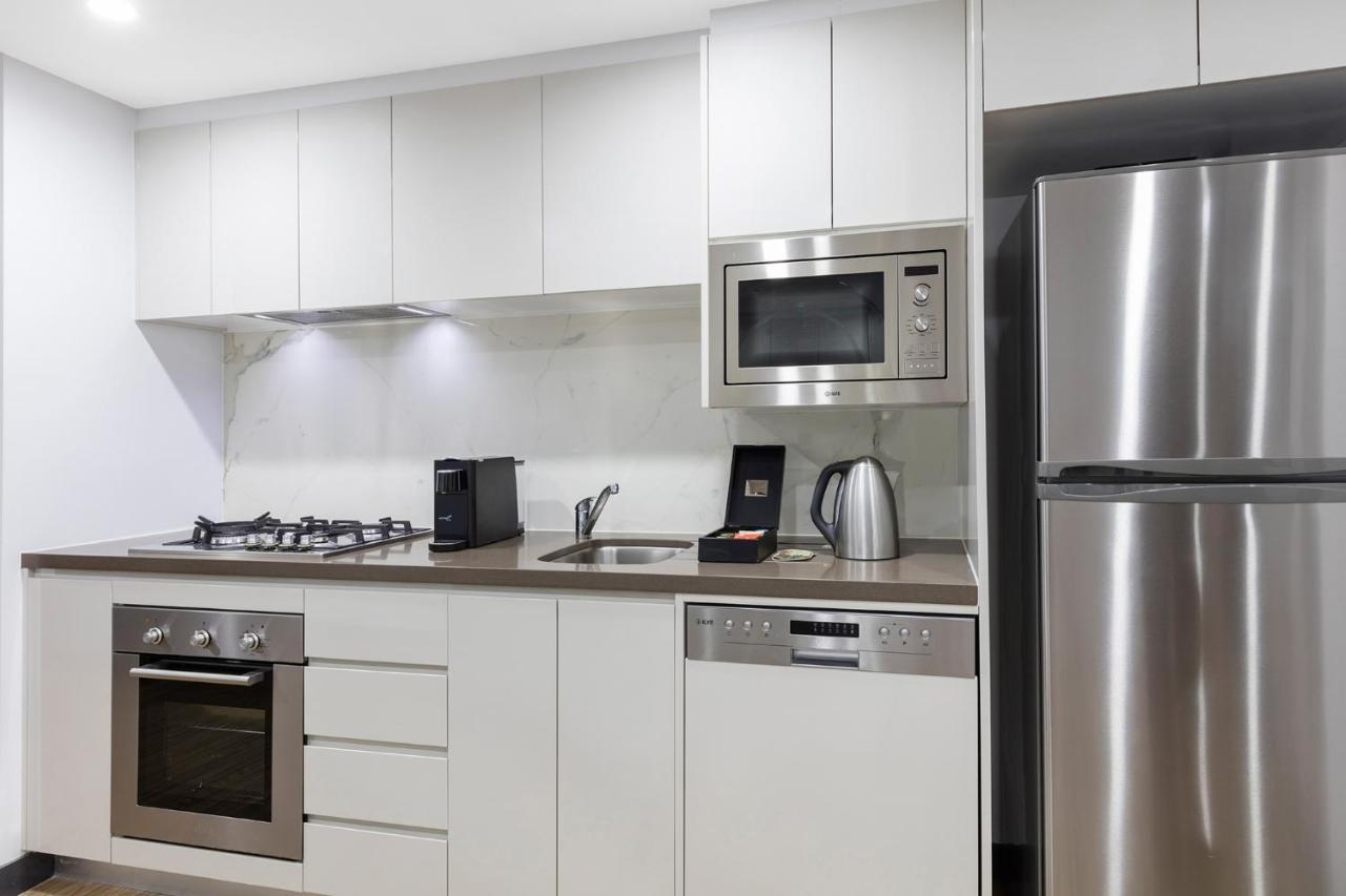 Meriton Suites Bondi Junction Sydney Kültér fotó The photo shows a modern kitchen with a sleek, minimalist design. It features white cabinetry and a countertop that blends with the overall aesthetic. 

Key elements include:

- A stainless steel refrigerator on the right.
- An oven with a gas cookto