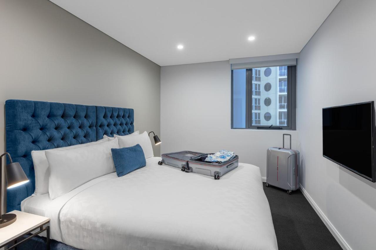 Meriton Suites Bondi Junction Sydney Kültér fotó The photo shows a modern hotel room featuring a neatly made bed with a blue tufted headboard. There is a small blue decorative pillow on the bed, and the bedding is white. Next to the bed, there is a nightstand with a lamp. A suitcase is partially op
