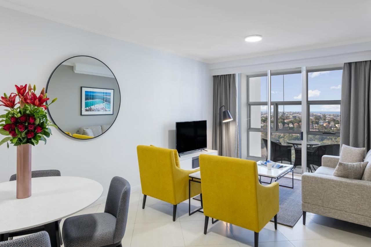 Meriton Suites Bondi Junction Sydney Kültér fotó The photo shows a modern living space with a stylish design. There are two bright yellow chairs facing a small glass table, and a comfortable beige sofa to the right. A TV is positioned on the wall in front of the chairs. To the left, there's a dinin