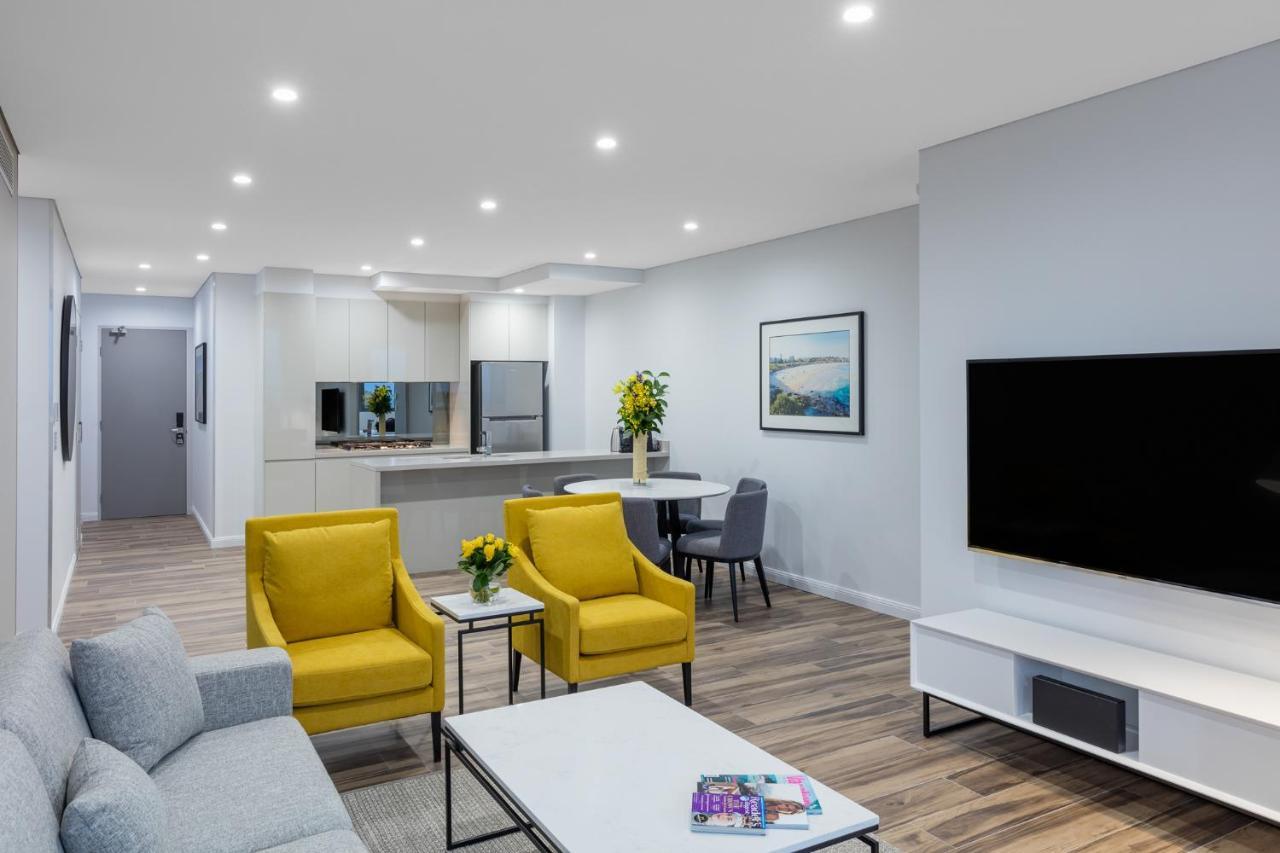 Meriton Suites Bondi Junction Sydney Kültér fotó The photo shows a modern living space that includes a cozy sitting area and a dining area. The sitting area features two bright yellow chairs and a light gray sofa, arranged around a coffee table. There are decorative items on the table, including fl