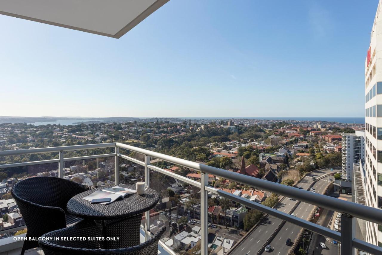 Meriton Suites Bondi Junction Sydney Kültér fotó The photo shows a balcony with a view over a cityscape that includes buildings and greenery, extending towards the horizon where the sea is visible. The balcony features a small round table and two black chairs, suggesting a space for relaxation or d