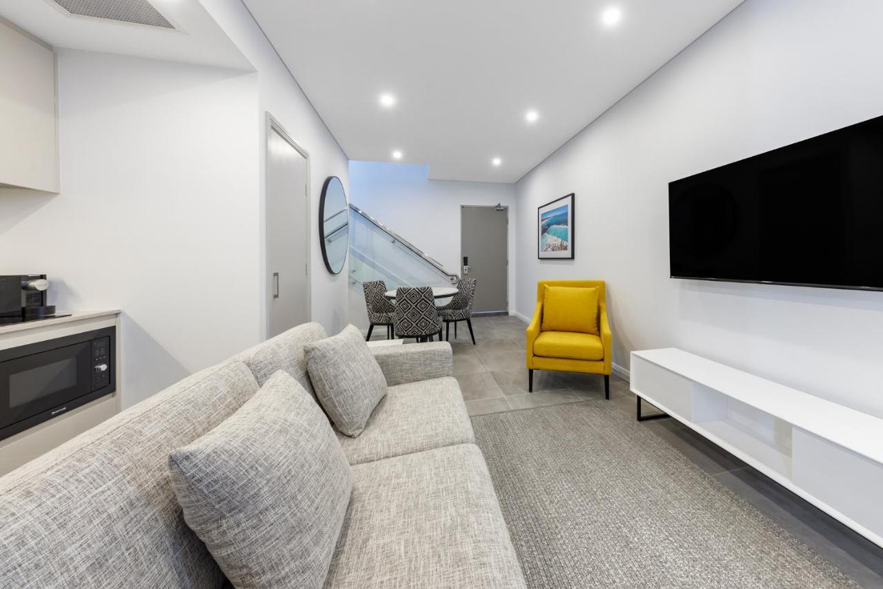 Meriton Suites Bondi Junction Sydney Kültér fotó The photo shows a modern and stylish living space. It features a light-colored sofa with cushions, positioned to face a wall-mounted flat-screen TV. There's a yellow accent chair nearby that adds a pop of color to the room. The décor is minimalistic,
