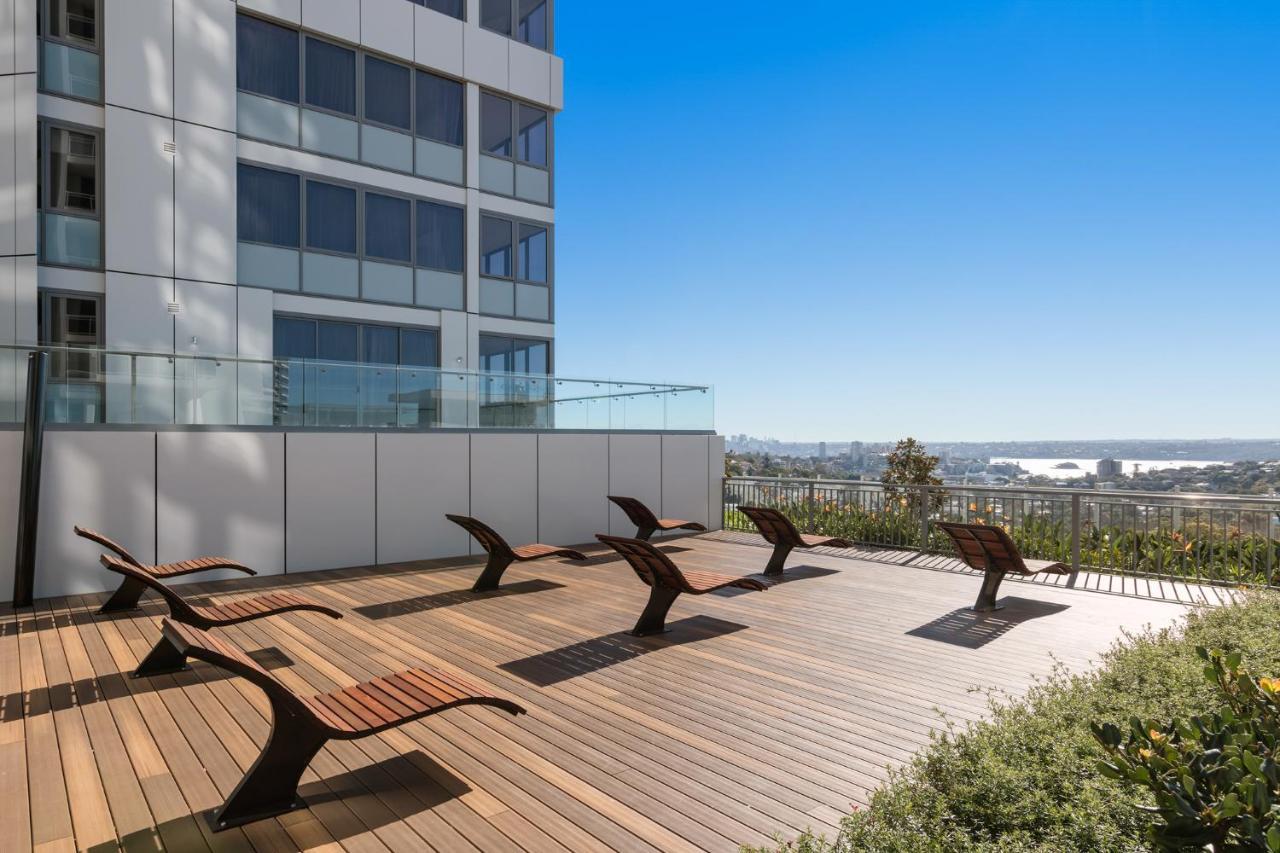 Meriton Suites Bondi Junction Sydney Kültér fotó The photo shows a rooftop or outdoor area with a wooden deck. There are several modern lounge chairs arranged neatly, facing a clear sky and a panoramic view of the surrounding landscape. In the background, there is a tall building with large windows