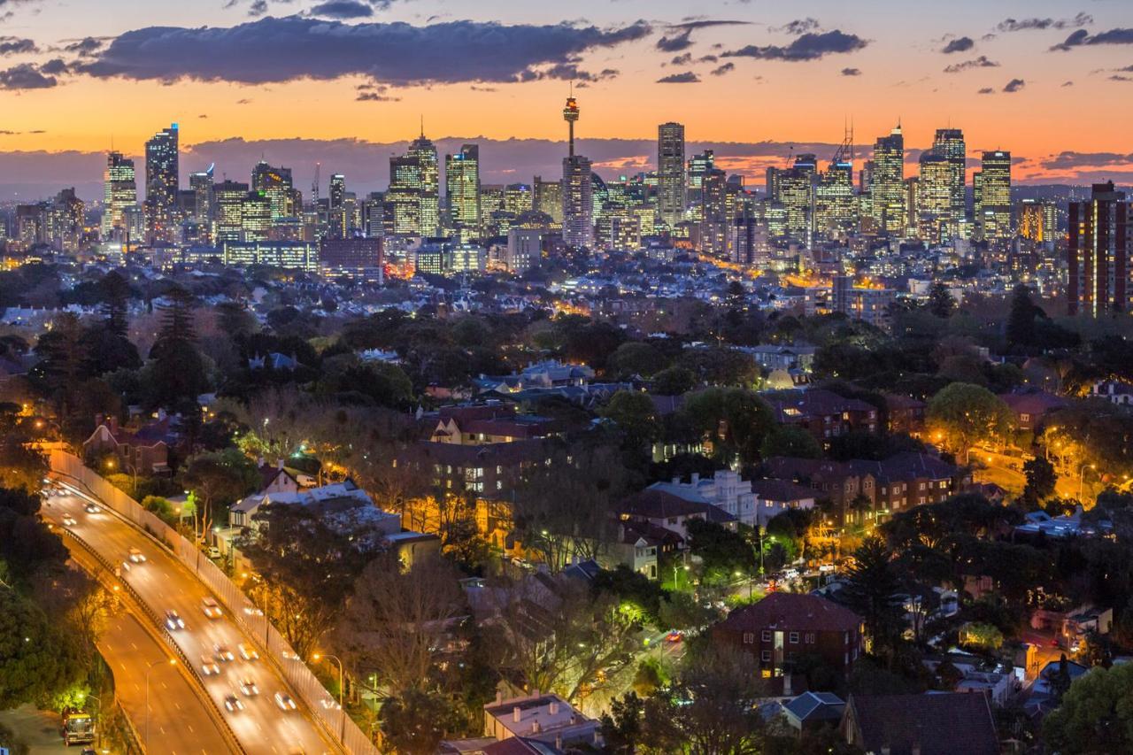 Meriton Suites Bondi Junction Sydney Kültér fotó The photo captures a stunning city skyline at dusk, featuring towering skyscrapers adorned with lights against a colorful sunset backdrop. In the foreground, a green area with trees and houses is visible, while a road with cars can be seen winding th