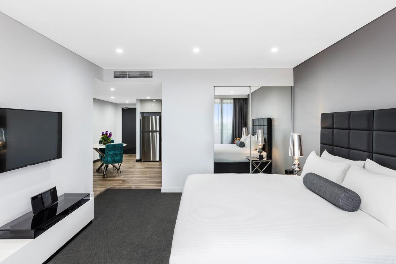 Meriton Suites Bondi Junction Sydney Kültér fotó The photo shows a modern hotel room. It features a large bed with a white covering and gray accent pillows. To the left, there's a flat-screen TV mounted on the wall above a sleek white cabinet. The flooring is a light wood tone, and a large mirror i