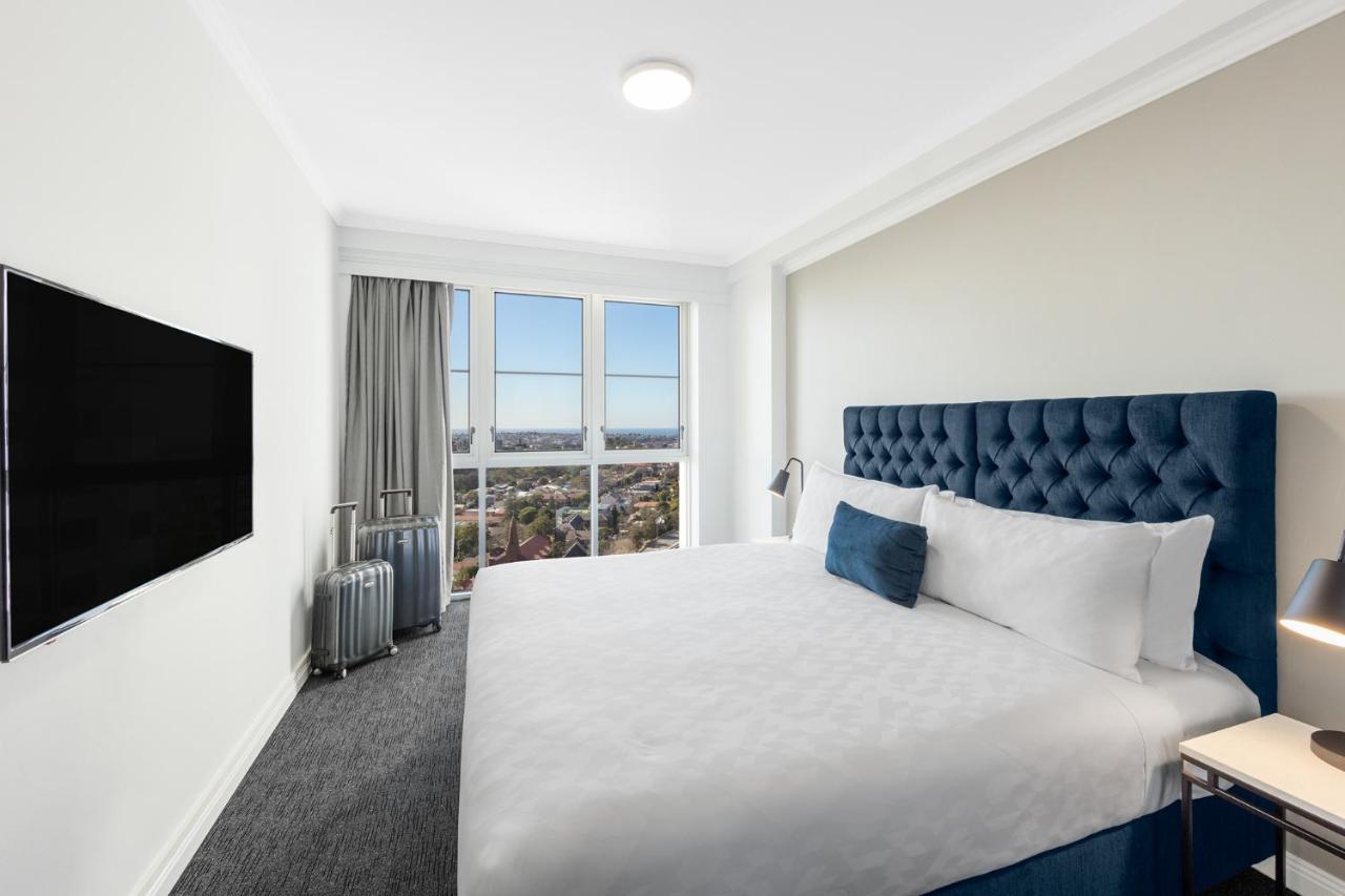 Meriton Suites Bondi Junction Sydney Kültér fotó The photo shows a modern hotel room with a large bed featuring a tufted blue headboard. There is a white duvet and a decorative pillow on the bed. A flat-screen television is mounted on the wall opposite the bed. Large windows are covered with gray c