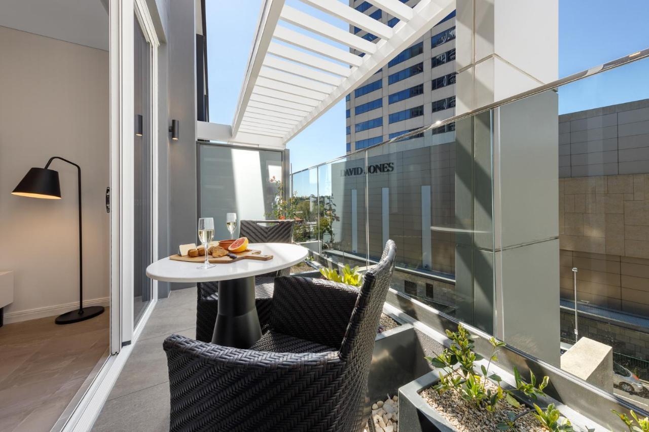 Meriton Suites Bondi Junction Sydney Kültér fotó The photo shows a modern balcony area with glass railings. It features a small round table set for two, with a plate of food and drinks. There are two stylish wicker chairs positioned around the table. The space is bright and airy, with sunlight stre
