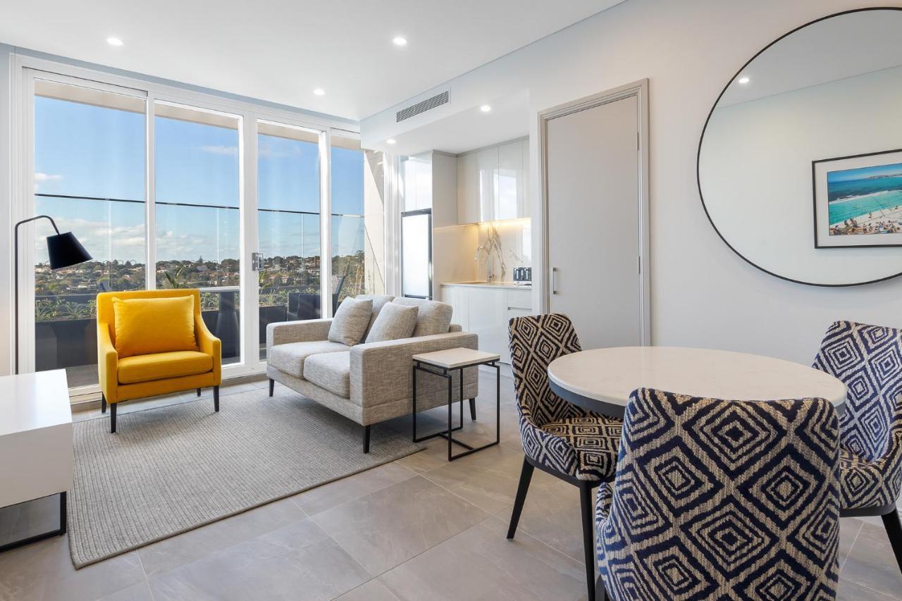 Meriton Suites Bondi Junction Sydney Kültér fotó The photo shows a modern and stylish living area. It features a comfortable sofa in a neutral color, accompanied by a bright yellow armchair that adds a pop of color. There's a round dining table with patterned chairs nearby. Large windows allow plen