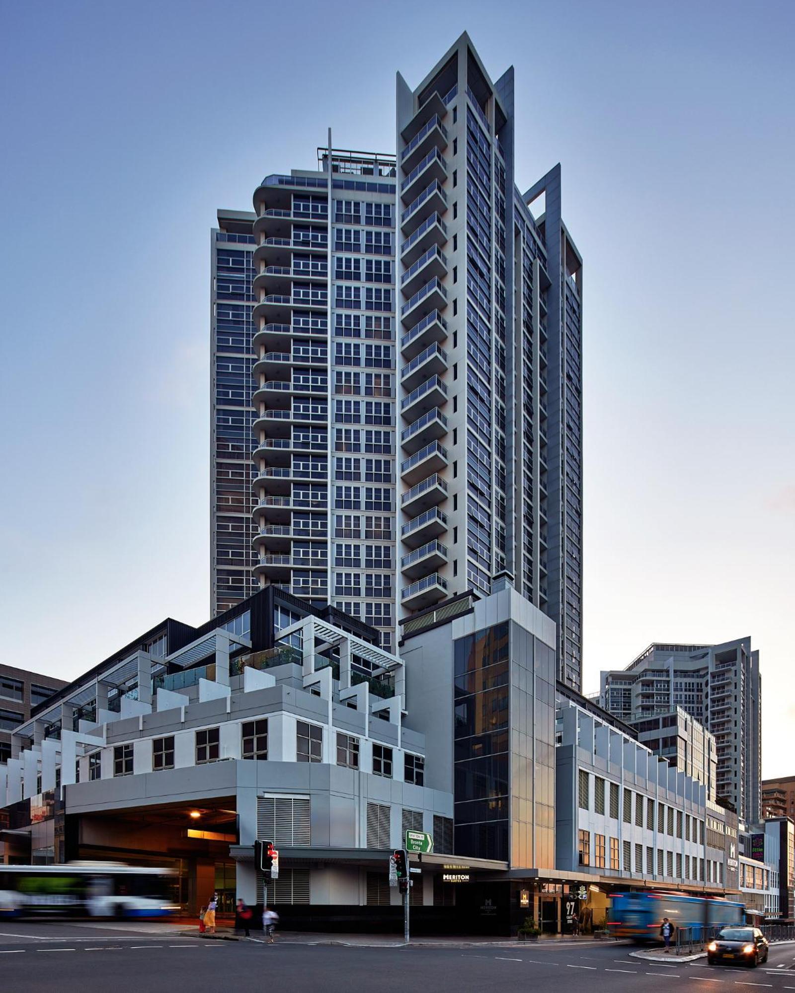Meriton Suites Bondi Junction Sydney Kültér fotó The image shows a modern high-rise building with a sleek design. The structure features a mix of glass and concrete elements, with multiple levels showcasing balconies. The building appears to be part of an urban environment, likely containing reside