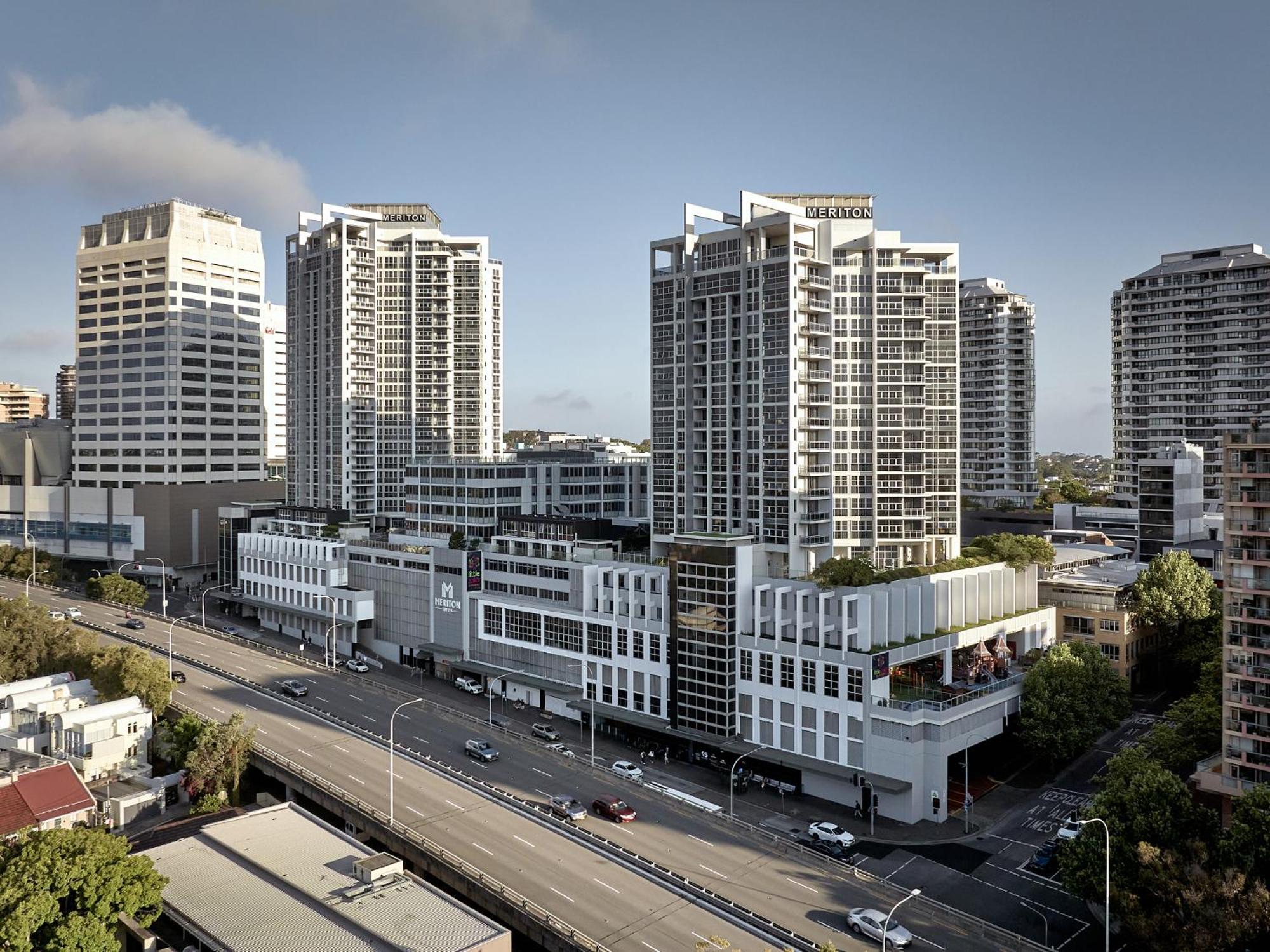 Meriton Suites Bondi Junction Sydney Kültér fotó The photo shows a cityscape featuring several tall modern buildings, likely apartments or office complexes. In the foreground, there is a road lined with cars, and the architecture of the buildings varies, with some having sleek glass facades and oth
