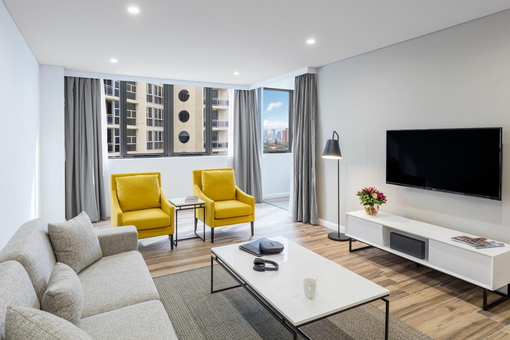Meriton Suites Bondi Junction Sydney Kültér fotó The photo shows a modern, well-lit living room. It features a light gray sofa with a textured fabric. There are two bright yellow armchairs positioned near a window, which allows natural light to enter the space. The room has light gray curtains and 